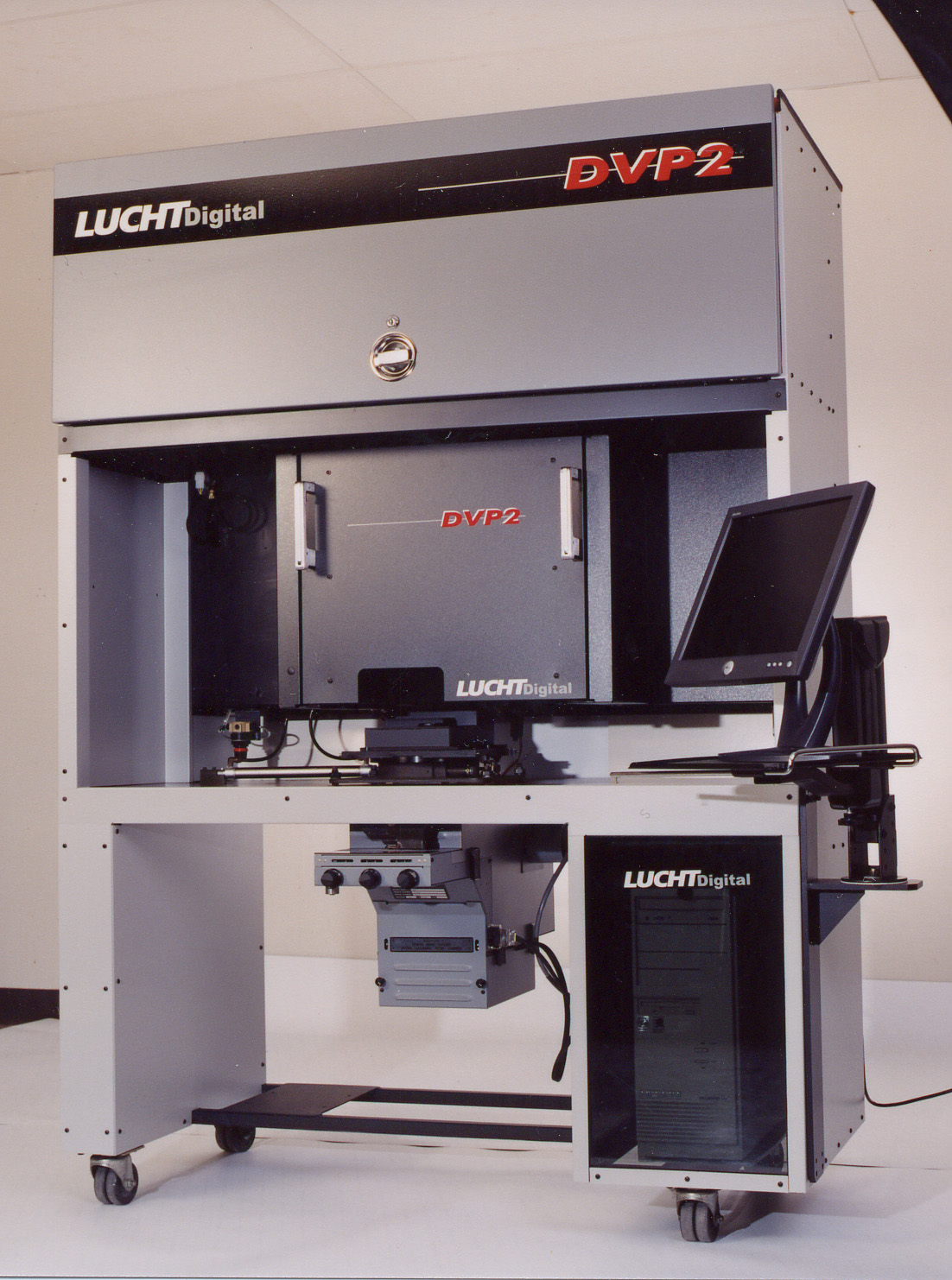DVP2 production cabinet, studio photograph — LUCHTdigital + DVP2 cabinet branding, lockable daylight canister door at the top, the patent-pending Sony 8-megapixel LCD-array print engine inside the central chassis, the touch-screen LCD on a swing arm at right, and the Dell-class controller PC in the LUCHTdigital-branded enclosure at the base.