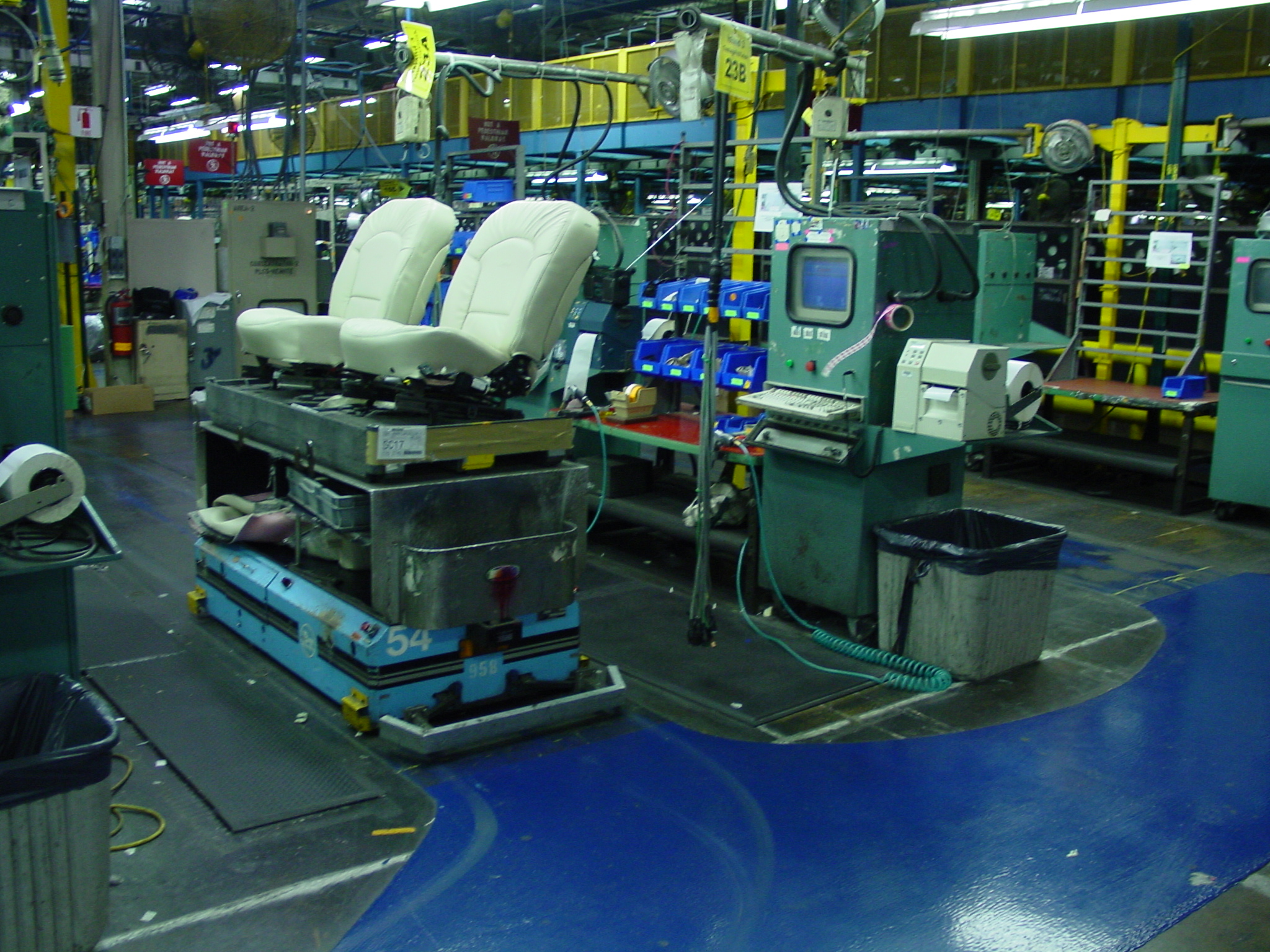 Ford Mustang seat assembly line in production — a rolling carrier with twin white-leather seats parked at one of the End-of-Line testers; original Motorola-era operator-display station visible at right